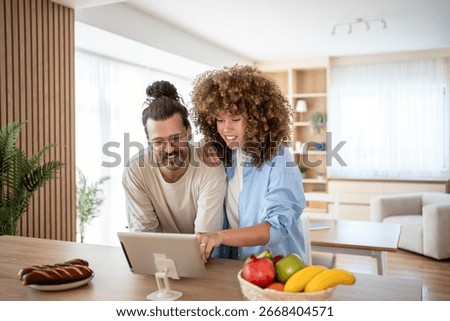 Smiling couple using digital tablet, browsing internet or watching movie, enjoying leisure time at home, healthy lifestyle with fresh fruits and bread in modern kitchen