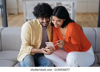 Smiling Couple Using Mobile Phone Shopping Online, Ordering Food Sitting At Home. Happy Relaxed African American Woman And Indian Man Holding Smartphone Communication, Looking At Digital Screen