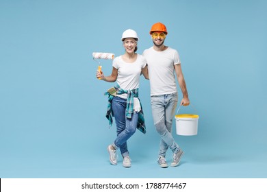 Smiling Couple Woman Man In Protective Helmet Hardhat Glasses Hold Paint Bucket, Paint Roller Isolated On Blue Background. Instruments Accessories For Renovation Apartment Room. Repair Home Concept