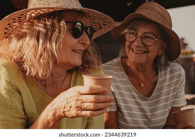 Smiling couple of women with hats make confidences while drink a coffee cup. Senior and middle aged female friends enjoying freedom and vacation in outdoors - Powered by Shutterstock