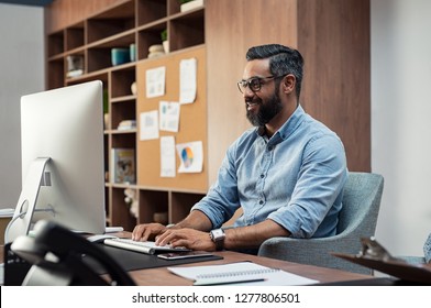 Smiling Creative Latin Business Man Typing On Desktop Computer In Office. Mature Middle Eastern Businessman Working At Modern Office Space. Happy Indian Man Wearing Eyeglasses Working On His Computer.