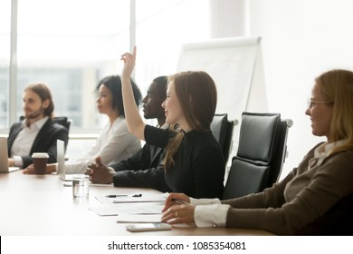 Smiling Curious Young Businesswoman Raising Hand At Multiracial Group Meeting Engaging In Offered Activity, Voting As Volunteer Or Asking Question At Corporate Business Training, Seminar Or Workshop