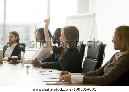 Smiling curious young businesswoman raising hand at multiracial group meeting engaging in offered activity, voting as volunteer or asking question at corporate business training, seminar or workshop