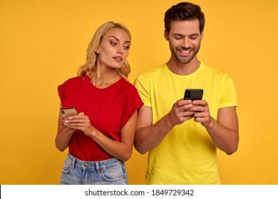 Smiling curious young couple friends guy girl in colored t-shirts posing isolated on yellow background studio. People lifestyle concept. Mock up copy space. Using mobile phones typing sms messages
