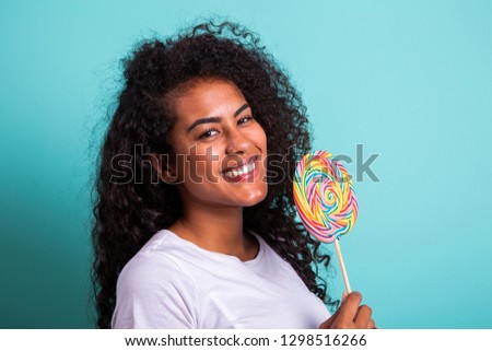 Smiling curly african girl in bright white t-shirt holding candy lollipop. Young cheerful woman hiding front face with lollipop at green studio background, copy space. Sweet life and confectionary
