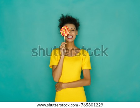 Smiling curly african girl in bright yellow dress holding candy lollipop. Young cheerful woman hiding out eye with lollipop at azur studio background, copy space. Sweet life and confectionary