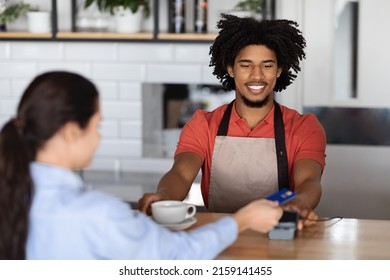 Smiling Curly Attractive Young Black Man Bartender In Apron At Bar Counter Gives Terminal To Lady Client With Credit Card In Cafe Interior. Order, Finance, Work And Small Business Due Covid-19