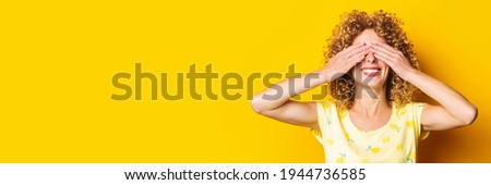 smiling curly girl covered her eyes with her palms on a yellow background. Banner