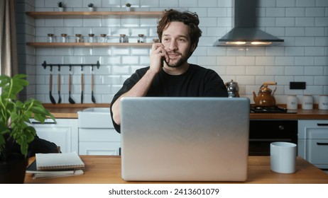 Smiling curly hair man sitting in the modern kitchen talking on cellphone while typing on laptop - Powered by Shutterstock