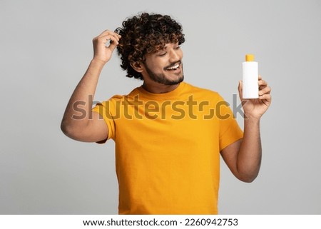 Smiling curly haired Indian man holding bottle with hair shampoo, looking at cosmetic product isolated on gray background, studio shot. Hair care concept