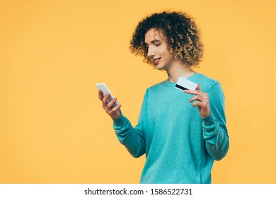 Smiling Curly Teenager Holding Smartphone And Credit Card Isolated On Yellow