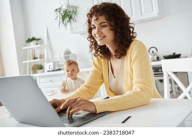 smiling curly woman typing on laptop while working remotely in kitchen near toddler daughter - Powered by Shutterstock