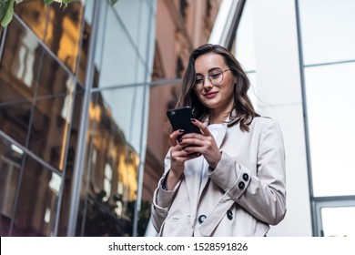 Smiling curly woman wearing trendy sunglasses walks down the central city street and uses her phone. Pretty summer woman in white jacket walks down the street looking at her mobile phone - Shutterstock ID 1528591826