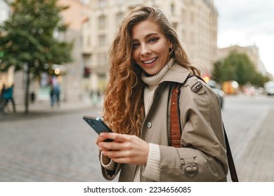 Smiling curly woman wearing warm coat walking down the street and using her phone
