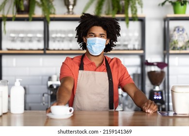 Smiling Curly Young Black Guy Bartender In Protective Mask And Apron Gives Cup Of Coffee To Client In Cafe Interior. Work And New Normal, Health Care During Covid-19 Outbreak, Startup And Business