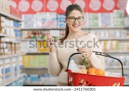 Smiling customer holding a shopping basket and showing a blank loyalty card at the supermarket, grocery loyalty program and discounts concept