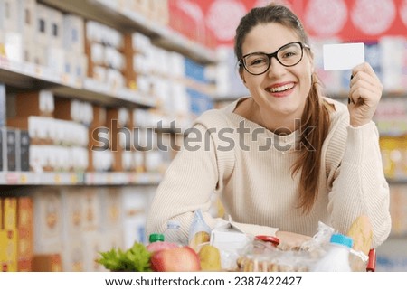 Smiling customer leaning on a full shopping cart and showing a blank loyalty card at the supermarket, grocery loyalty program and discounts concept