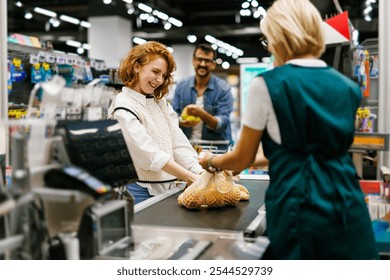 Smiling customer placing groceries on conveyor belt while friendly cashier scanning items at checkout counter in modern supermarket - Powered by Shutterstock