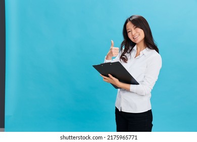 Smiling Cute Asian Businesswoman In Classic Dress Code Holds Folder Tablet With Application Form Show Thumb Up Gesture Posing Isolated On Blue Background. Cool Business Offer. Job Interview Concept