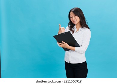 Smiling Cute Asian Businesswoman In Classic Dress Code Holds Folder Tablet With Application Form Show Thumb Up Gesture Posing Isolated On Blue Background. Cool Business Offer. Job Interview Concept