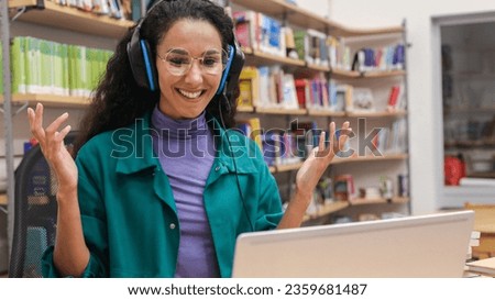 Smiling cute Eastern girl in a glasses in the library talking on a video call using headphones Library worker freelance intern having a pleasant video conversation gesticulating looking at the screen.