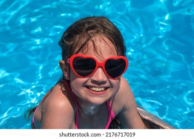 Smiling Cute Kid Girl In Heart Shaped Sunglasses In Swimming Pool On Sunny Day. Childhood Summer Vacation Concept. Happy Child Playing. Healthy Outdoor Sport Activity For Children. Kids Beach Fun