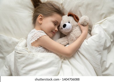 Smiling cute little girl hug teddy bear sleep peacefully in white bed relaxing on soft pillow, calm preschooler child enjoy daydream with plush toy taking nap under warm fluffy duvet at home