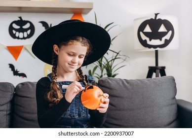 Smiling cute little girl preparing for Halloween  painting drawing face pumpkin while sitting cozy sofa in living room at home  smiling child making jack  o  lantern  Holiday decoration concept