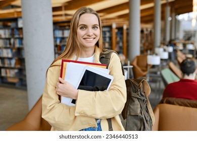 Smiling cute pretty blond girl, positive female teenage high school student holding backpack looking at camera standing in modern university or college campus library, portrait.
