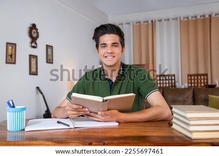 Smiling cute teenage boy studying with books - indoors stock photo. Royalty free image smart boy student teenage boy, sitting on chair, studying with books, at home - smiling face