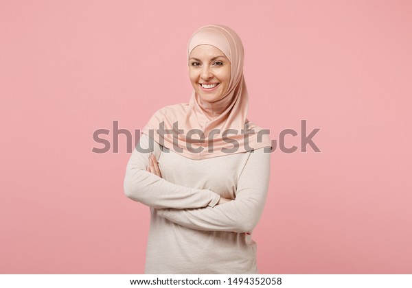 ピンクの背景にスタジオでポートするヒジャブの軽い服を着た 笑顔の若いアラビア人のイスラム教徒の女性 人々は宗教的なイスラムのライフスタイルのコンセプトを持つ コピー用スペースのモックアップ 手を組んで の写真素材 今すぐ編集