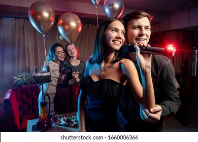 Smiling Cute Young Caucasian Couple Spend Free Time, Celebrating In Karaoke Bar, Man And Woman In Black Wear Singing Together In Microphone, Look Side