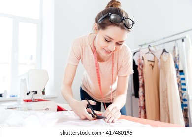 Smiling cute young woman seamstress standing and cutting white fabric with scissors