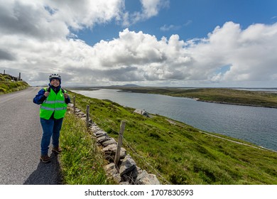 Smiling cyclist pausing on Sky Road coastal road, looking at camera, Connemara coast with blue sky and clouds in background, reflective vest and helmet, sunny day in Clifden, County Galway, Ireland - Powered by Shutterstock