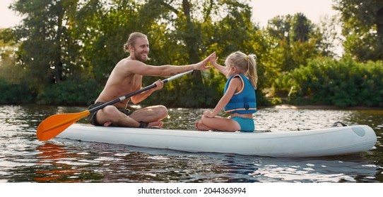 Smiling dad giving high five to his little daughter while relaxing, sup surfing on a river surrounded by the beautiful nature on a summer day. Sup board, travel, leisure concept