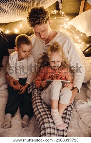 Smiling dad and his cute kids wearing pajamas watching cartoons on digital tablet while sitting in handmade tent in children's room