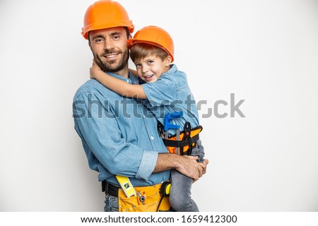 Smiling dad is holding boy on hands and hugging while they are wearing building helmets and belts with hand tools. Isolated on white background Foto d'archivio © 