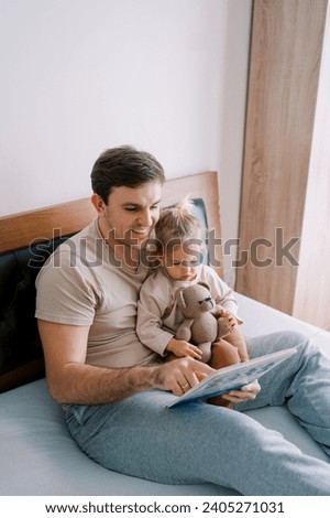 Smiling dad reading a book to a little girl sitting with a teddy bear on his lap on the bed