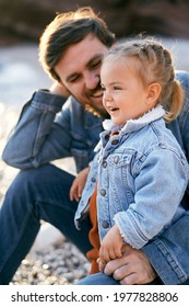 Smiling Dad Is Sitting Next To A Little Laughing Girl On A Pebble Beach. Close-up