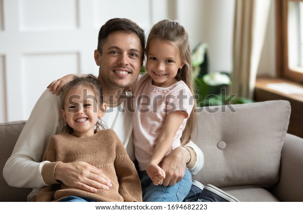 Smiling dad uncle holding on lap little\
children, looking at camera. Happy young single man relaxing on\
comfortable couch with adorable two daughters in living room,\
joyful family of three\
portrait.