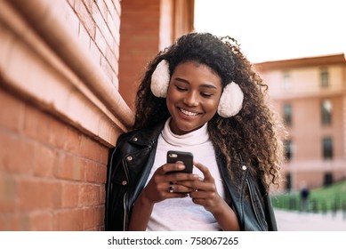 Smiling Dark Skinned Student Woman With Phone Leaning On Wall.