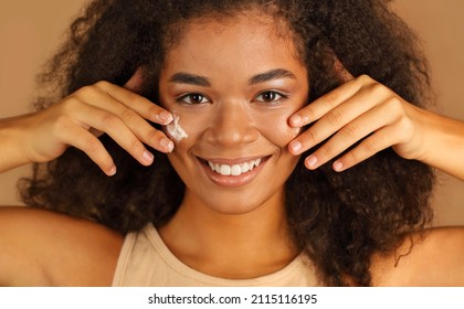 Smiling Dark Skinned Woman With Curly Afro Hair Applies Face Cream On Cheeks With Both Hands, Poses Over Beige Studio Wall Background, Uses Cosmetic Product. Skin Care And Beauty Treatments