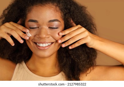 Smiling Dark Skinned Woman With Curly Afro Hair Applies Face Cream On Cheeks With Both Hands, Poses Over Beige Studio Wall Background, Uses Cosmetic Product. Skin Care And Beauty Treatments