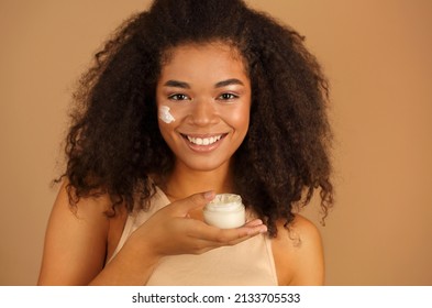 Smiling Dark Skinned Woman With Curly Afro Hair Applies Face Cream On Cheeks With Both Hands, Poses Over Beige Studio Wall Background, Uses Cosmetic Product. Skin Care And Beauty Treatments