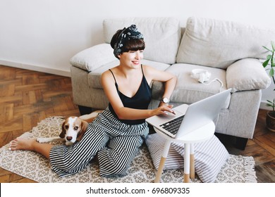 Smiling Dark-haired Lady In Trendy Striped Pants Working With Computer While Her Beagle Dog Lying Beside.  Excited Girl With Ribbon In Hair Looking At Laptop Screen Sitting On Carpet With Puppy.