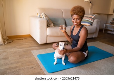 Smiling Dark-skinned Beautiful Young Woman Drinking Water From The Glass Sitting At Home In Living Room On Yoga Mat. Break After Exercises. Morning Routine. Petting Small Dog Jack Russell Terrier