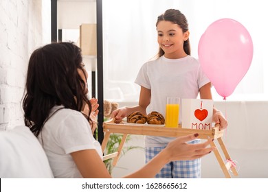 Smiling Daughter Holding Tray With Mothers Day Card With Heart Sign And Mom Lettering Near Mother Sitting In Bed