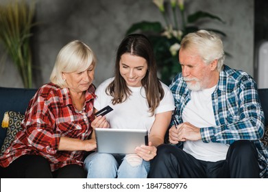 Smiling Daughter Sitting At Home On The Sofa With Her Elderly Parents And Teaching Them Online Shopping And Paying Utility Bills Using A Laptop And A Bank Card