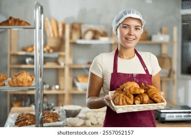 Smiling dedicated young female baker holding tray of golden freshly baked croissants, proudly offering freshly baked goods in cozy private bakery environment - Powered by Shutterstock