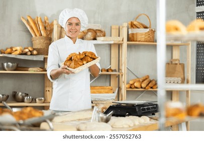 Smiling dedicated young female baker holding tray of golden freshly baked croissants, proudly offering freshly baked goods in cozy private bakery environment - Powered by Shutterstock